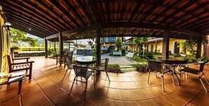 An outdoor patio area with tiled flooring, metal chairs, and tables. There is a covered section with a wooden roof and hanging plants. - Pousada do Sol (Ubatuba)