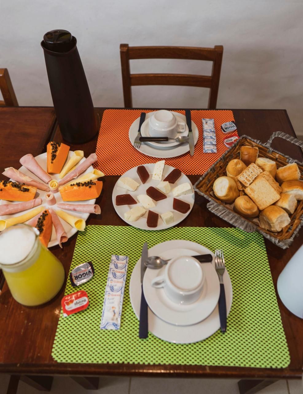 Une table dressée pour le petit-déjeuner, avec des assiettes garnies d’un assortiment de fromages, de charcuteries et de pain, ainsi que des condiments et un thermos.