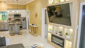 A modern living room with a large flat-screen TV, a fireplace, and a dining area with bar stools. - Cozy Luxury Hideaway With Free Wifi,Parking,Swimming Pool&Jacuzzi Near Negril 1b (Green Island)