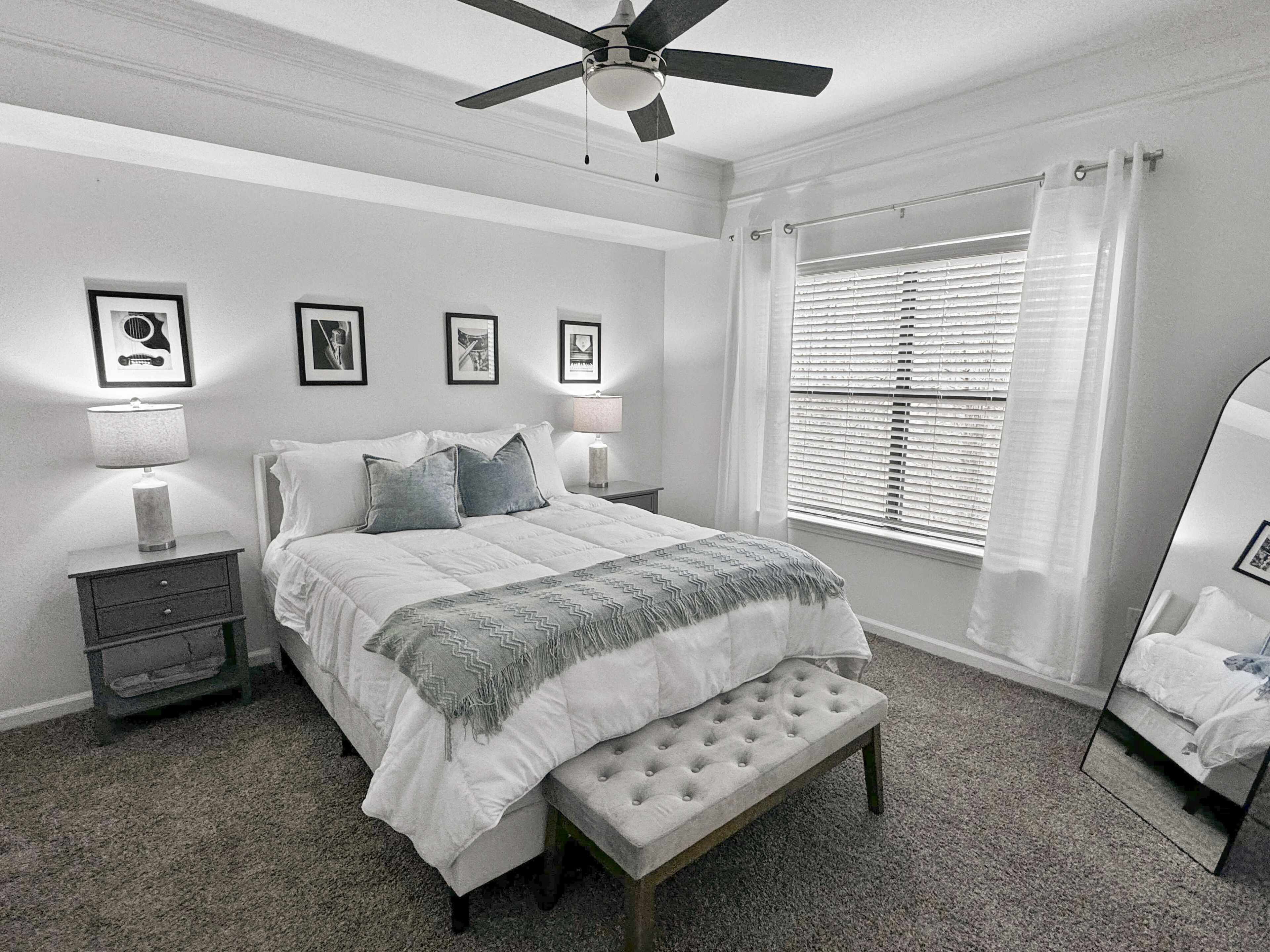 Un dormitorio con cama, ventilador de techo, ventana con persianas e imágenes enmarcadas en la pared.