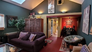 Ein Wohnzimmer mit einem violetten Sofa, einem grünen Sessel und einem roten Teppich. Es gibt ein Holzschränkchen mit Dekoration und einer Pflanze.