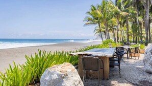 Eine Terrasse am Strand mit Tisch und Stühlen, umgeben von Palmen und Blick auf den Ozean.