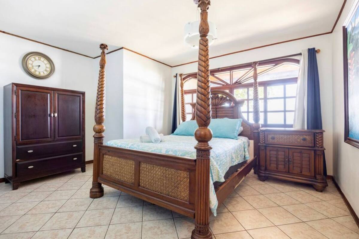 Ein Bett mit Baldachin, dessen Gestell aus Holz gefertigt ist und ein gewebtes Kopfende besitzt, ein dunkler Holzschrank sowie eine Wanduhr.