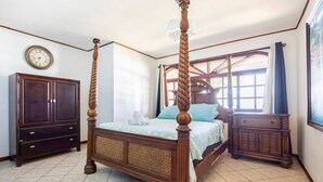 Ein Bett mit Baldachin, dessen Gestell aus Holz gefertigt ist und ein gewebtes Kopfende besitzt, ein dunkler Holzschrank sowie eine Wanduhr.