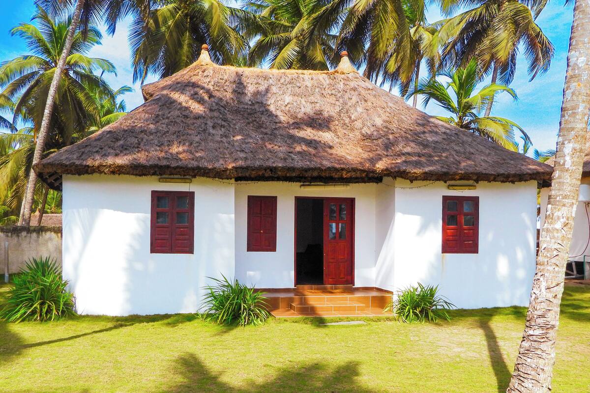 A white building with a thatched roof, red doors, and windows, surrounded by palm trees and grass.