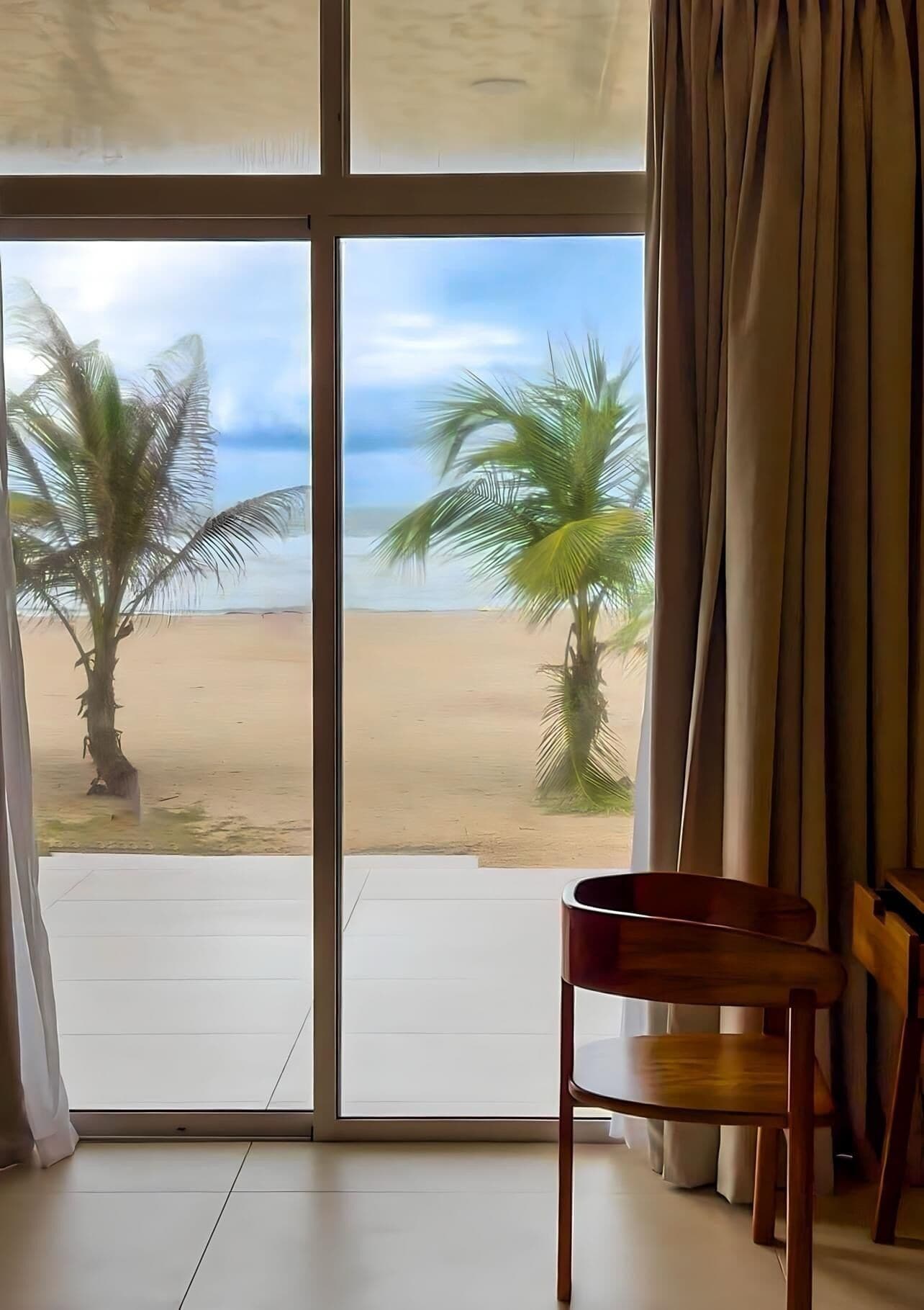 A room with a large sliding glass door leading to a beach view, featuring palm trees and a sandy shore.