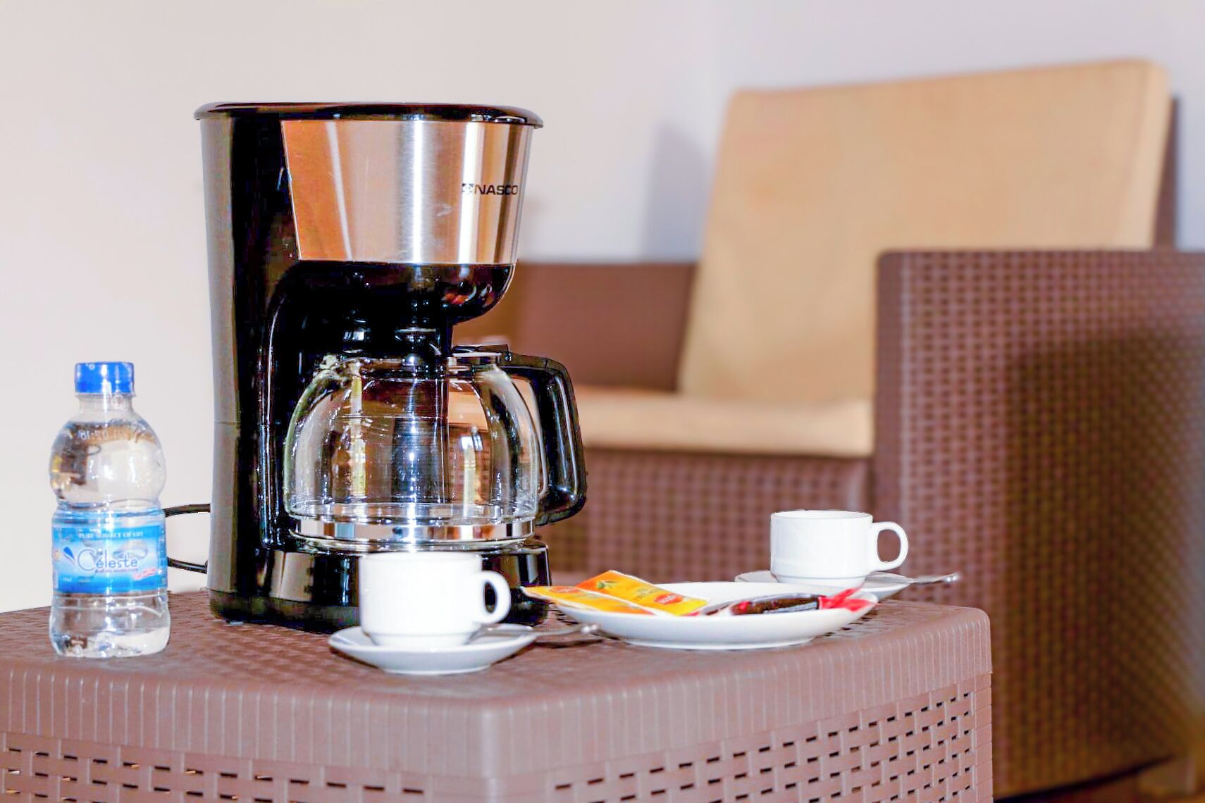 Una cafetera, una ampolla d'aigua, una tassa i un plat amb menjar sobre una taula petita en una habitació amb una cadira.