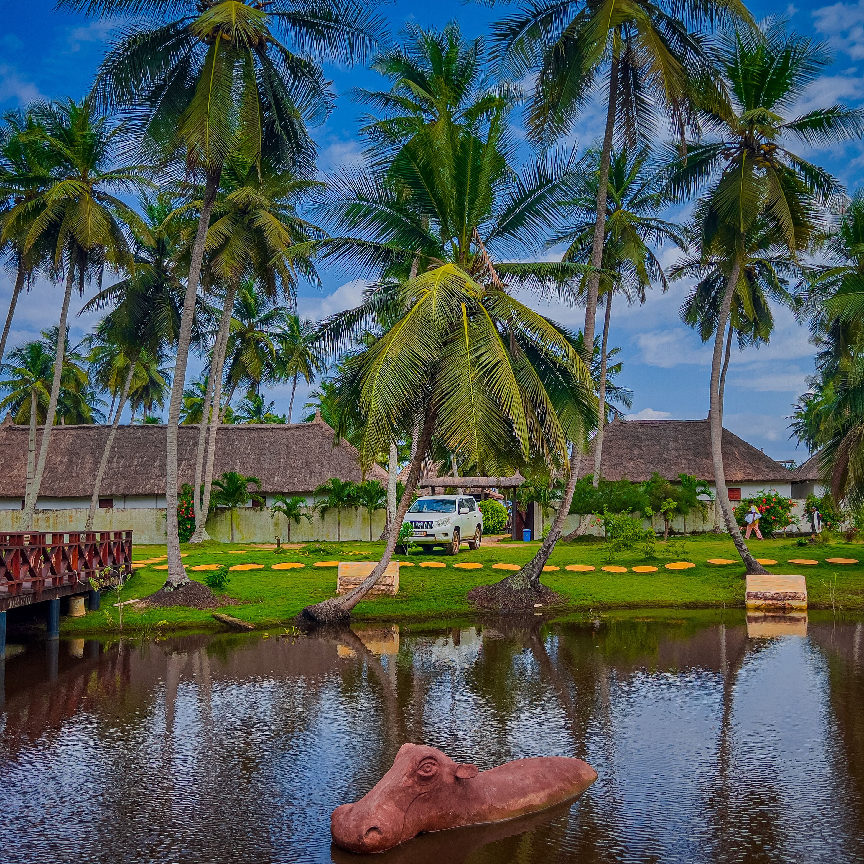 Um resort tropical com palmeiras, um edifício com telhado de palha e um pequeno lago com uma escultura.