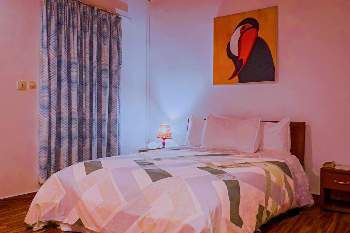 A bedroom with a bed, a nightstand, a lamp, a painting, and a window with curtains.