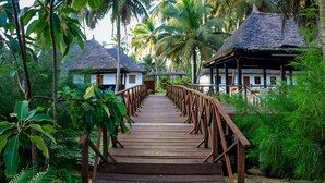 A wooden bridge leading to a thatched-roof building surrounded by lush greenery. - Sabbat Plage (Mohamé)