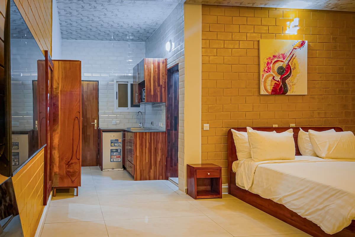 A bedroom with a bed, bedside tables, and a nightstand. There is a small wooden cabinet and a refrigerator in the adjacent kitchen area.