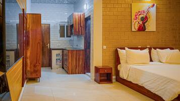 Un dormitorio con cama, mesitas de noche y mesilla auxiliar. Hay un pequeño armario de madera y un refrigerador en la cocina contigua.