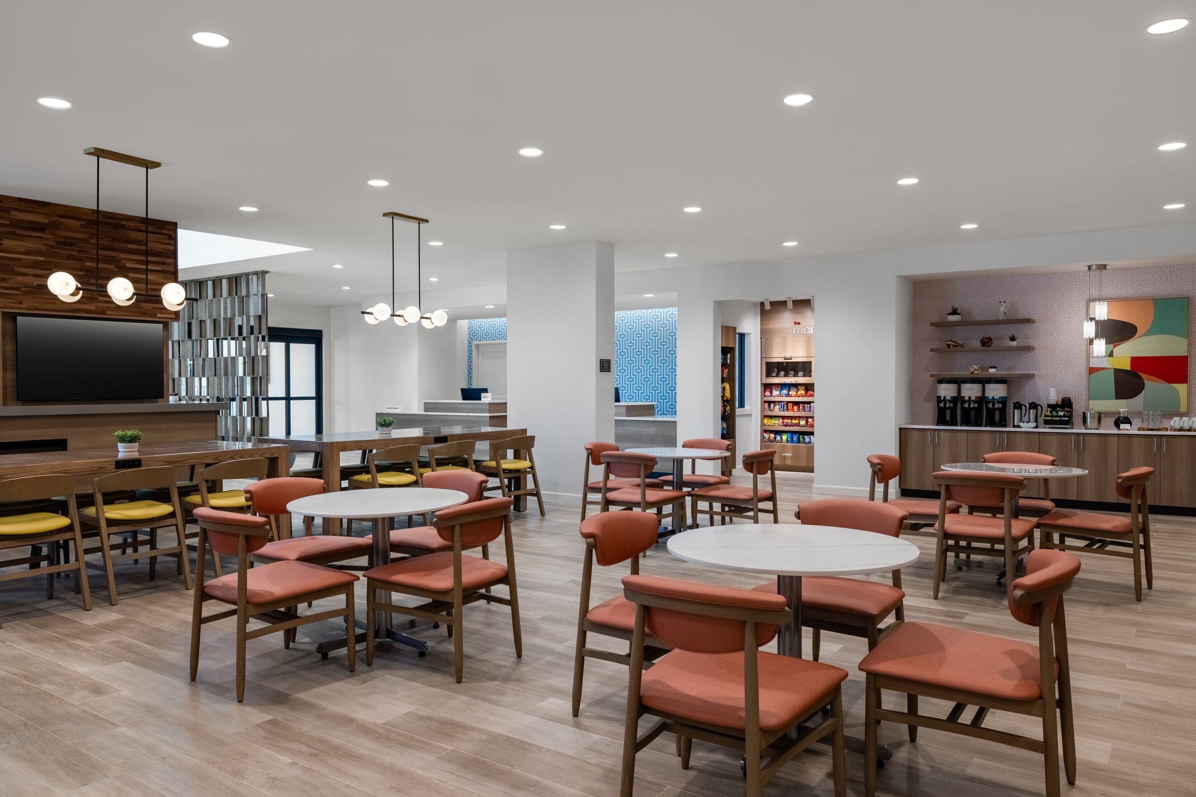 A modern dining area with round tables and orange chairs, a kitchen area with a coffee machine, and a large TV screen.