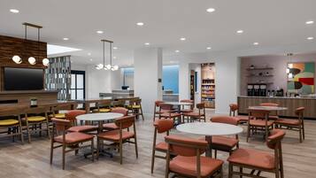 A modern dining area with round tables and orange chairs, a kitchen area with a coffee machine, and a large TV screen.