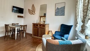 A living room with a TV, wooden dining table, blue armchair, and a cabinet with decorative items. - Belle Échappée, Expérience Unique, Alliant Charme Historique, Commodités Moderne (Le Crotoy)
