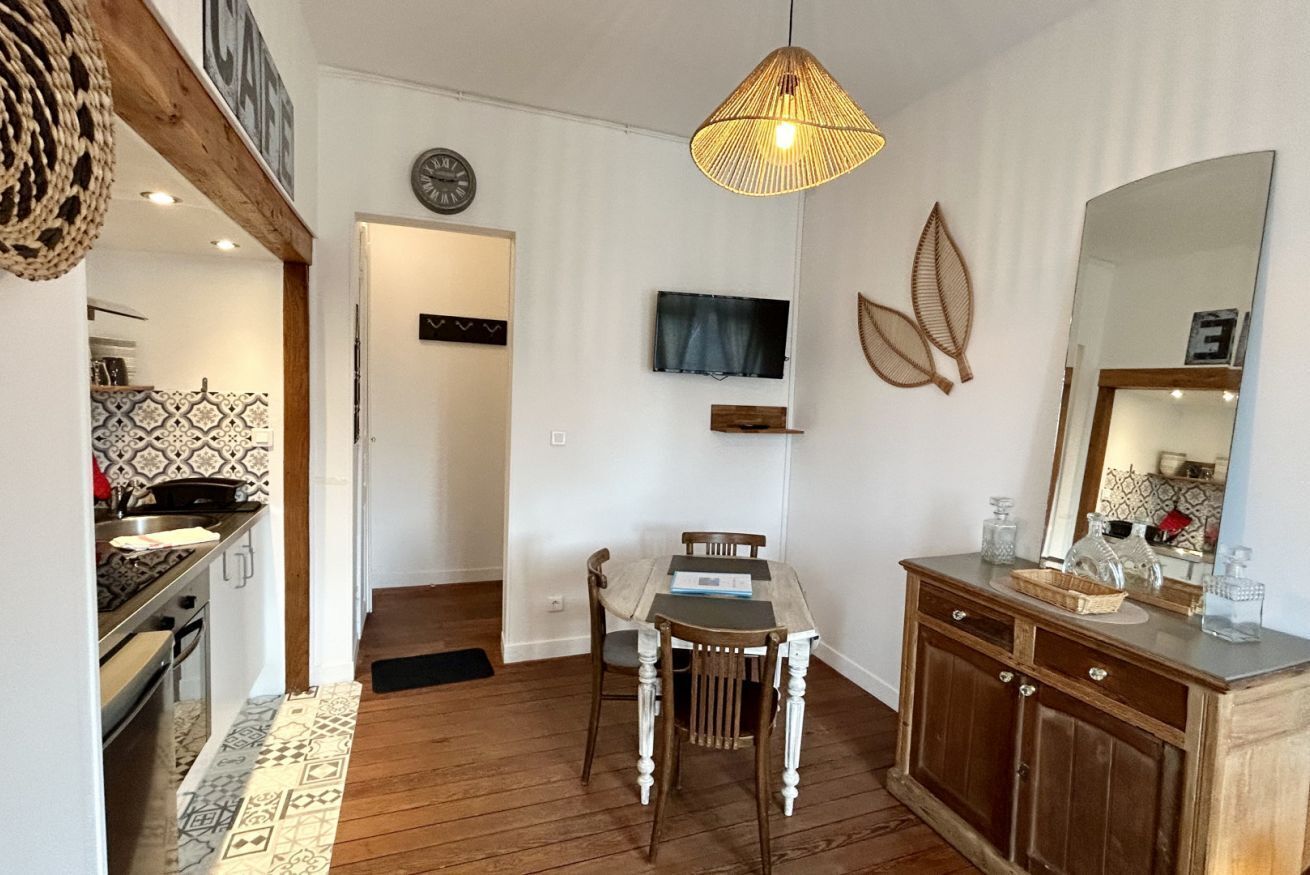 Un coin repas avec une table et des chaises en bois, un luminaire suspendu, un téléviseur fixé au mur et une cuisine visible au fond.