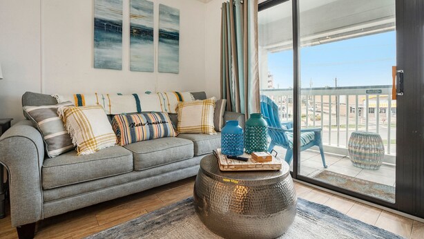 A living room with a grey sofa, decorative pillows, a round ottoman, and a view through a glass door to the outside.