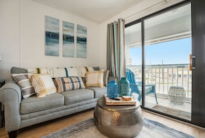 A living room with a grey sofa, decorative pillows, a round ottoman, and a view through a glass door to the outside. - Affordable cozy Galveston condo with beautiful ocean views, perfect getaway! (Galveston)