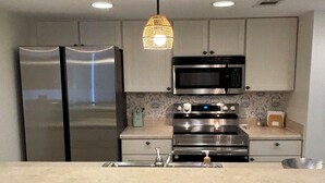 Une cuisine équipée d’un réfrigérateur, d’un four à micro-ondes et d’un four en acier inoxydable, un coin bar avec des chaises en osier et un luminaire suspendu.
