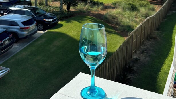 Un verre contenant un liquide bleu est posé sur un balcon donnant sur une plage où l’on aperçoit des voitures garées et des palmiers.