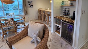 Un vaste espace de vie comprenant un canapé en osier, une table à manger et des chaises. On aperçoit également un coin cuisine avec un réfrigérateur et des étagères.