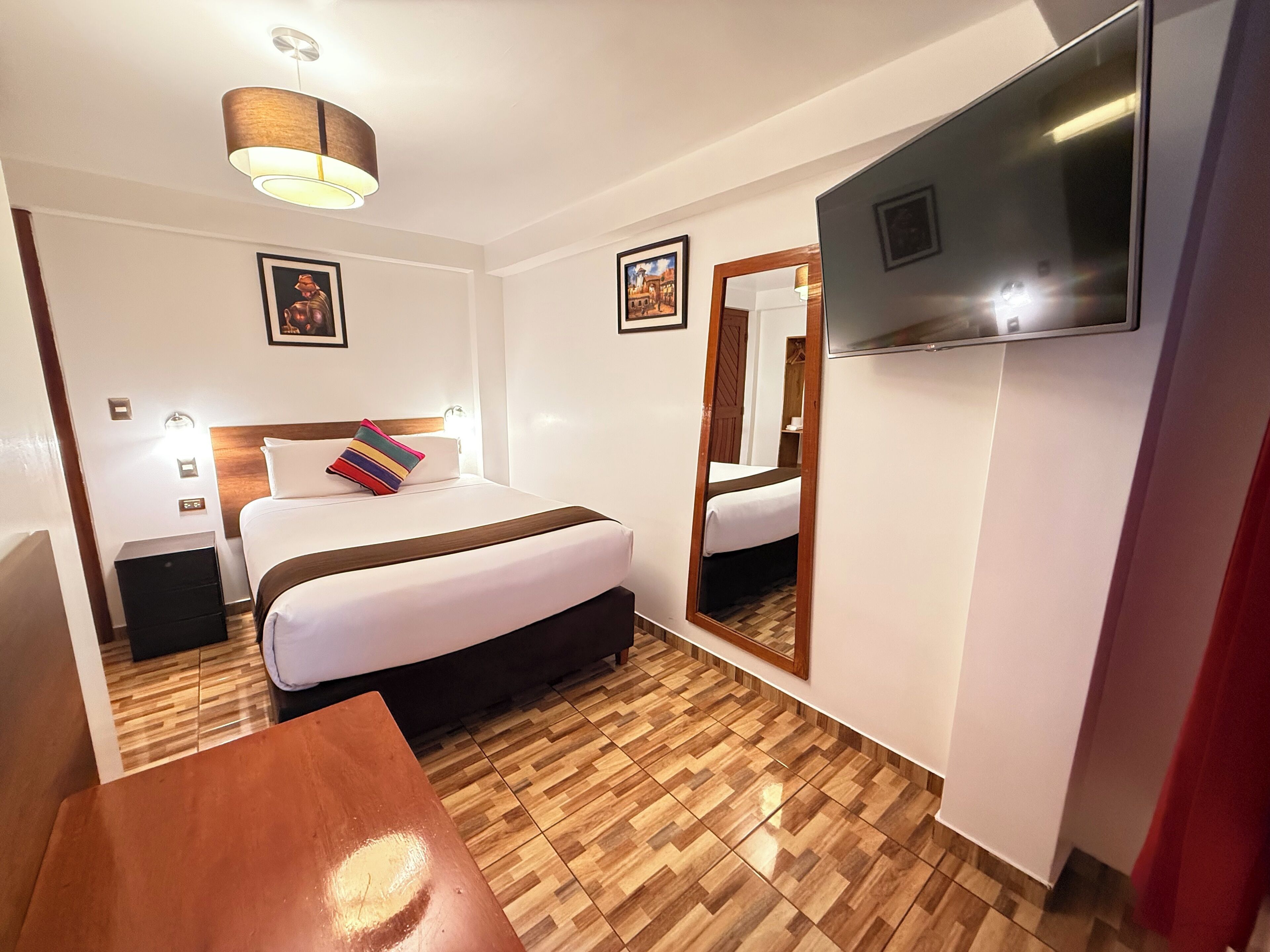 A hotel room with a bed, bedside table, lamp, TV, and wooden flooring.