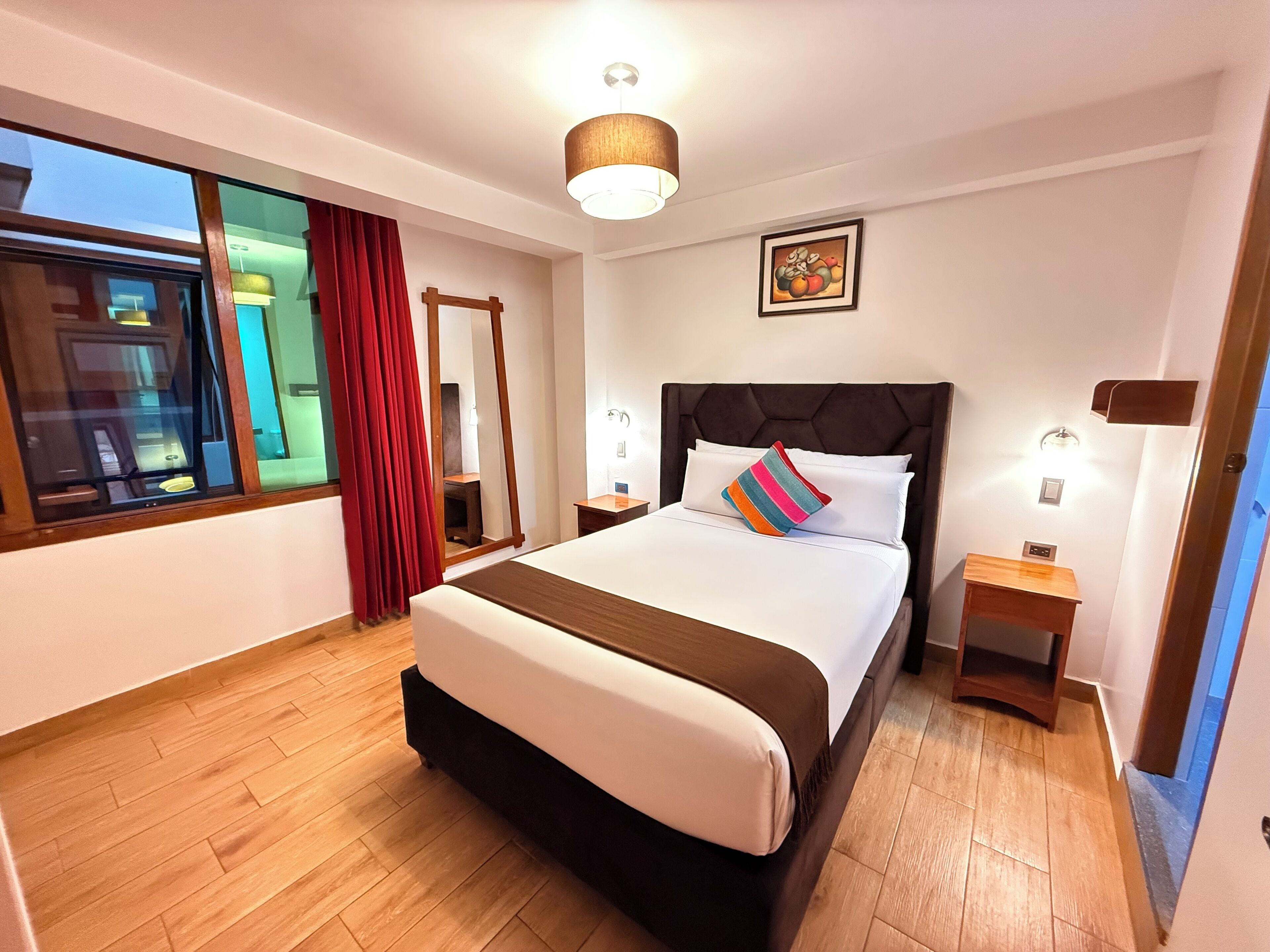 A hotel room with a large bed, wooden flooring, a window with curtains, a bedside table, and a wall-mounted light fixture.