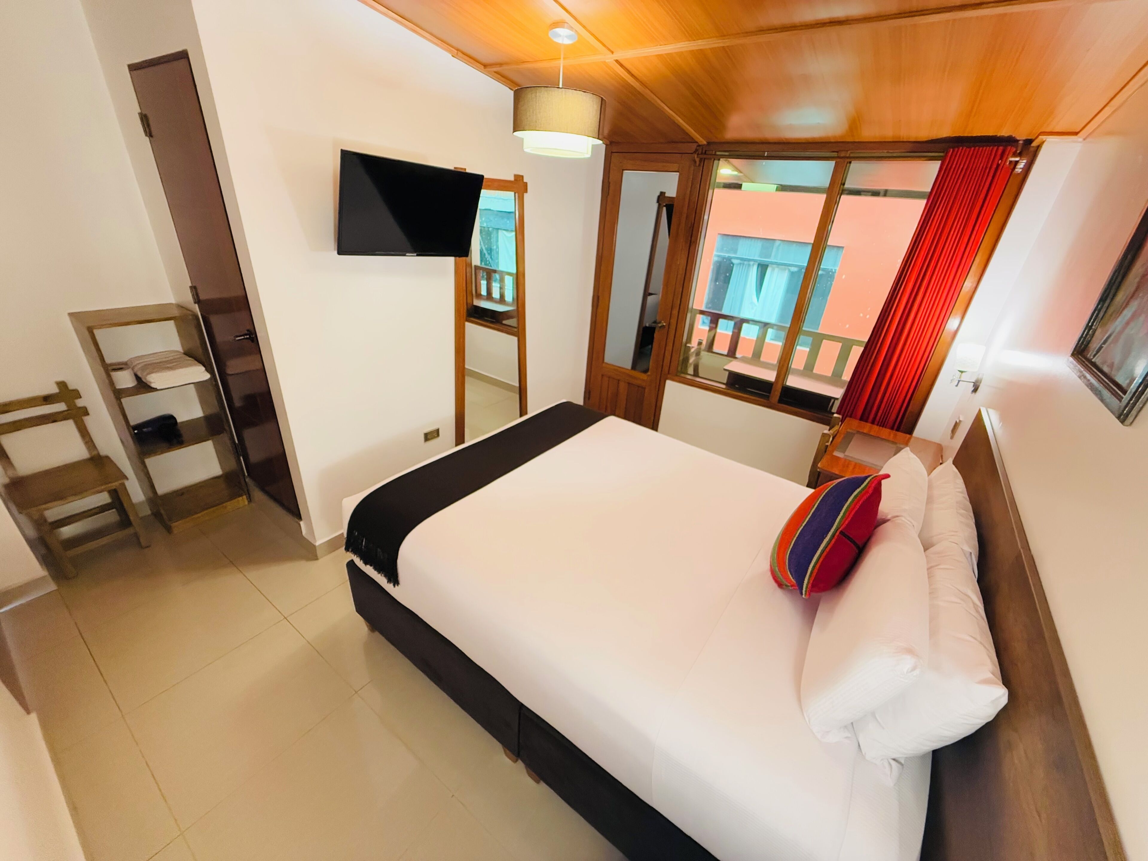 Habitación de hotel con cama, televisión montada en la pared, una silla de madera, una mesita y una ventana con cortinas.