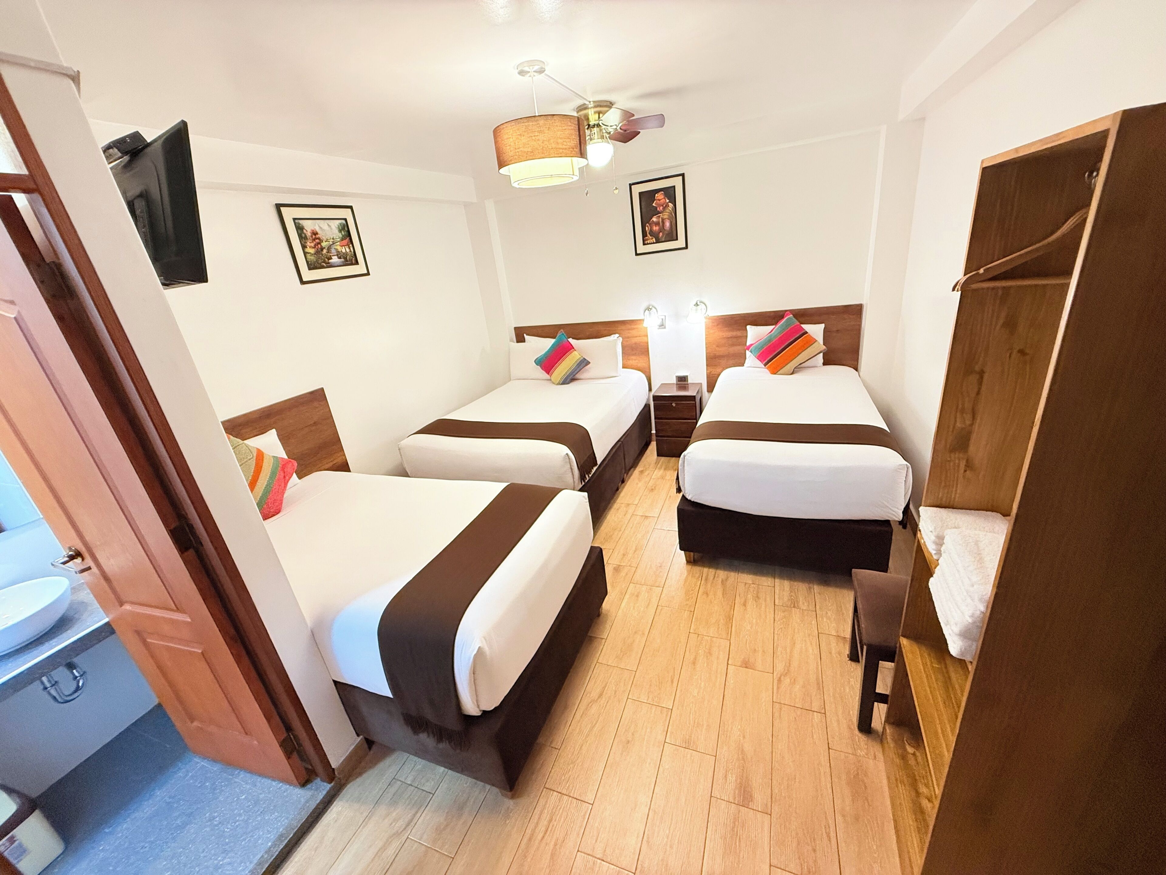 A hotel room with three beds, a wooden wardrobe, a ceiling fan, and a nightstand with a lamp.