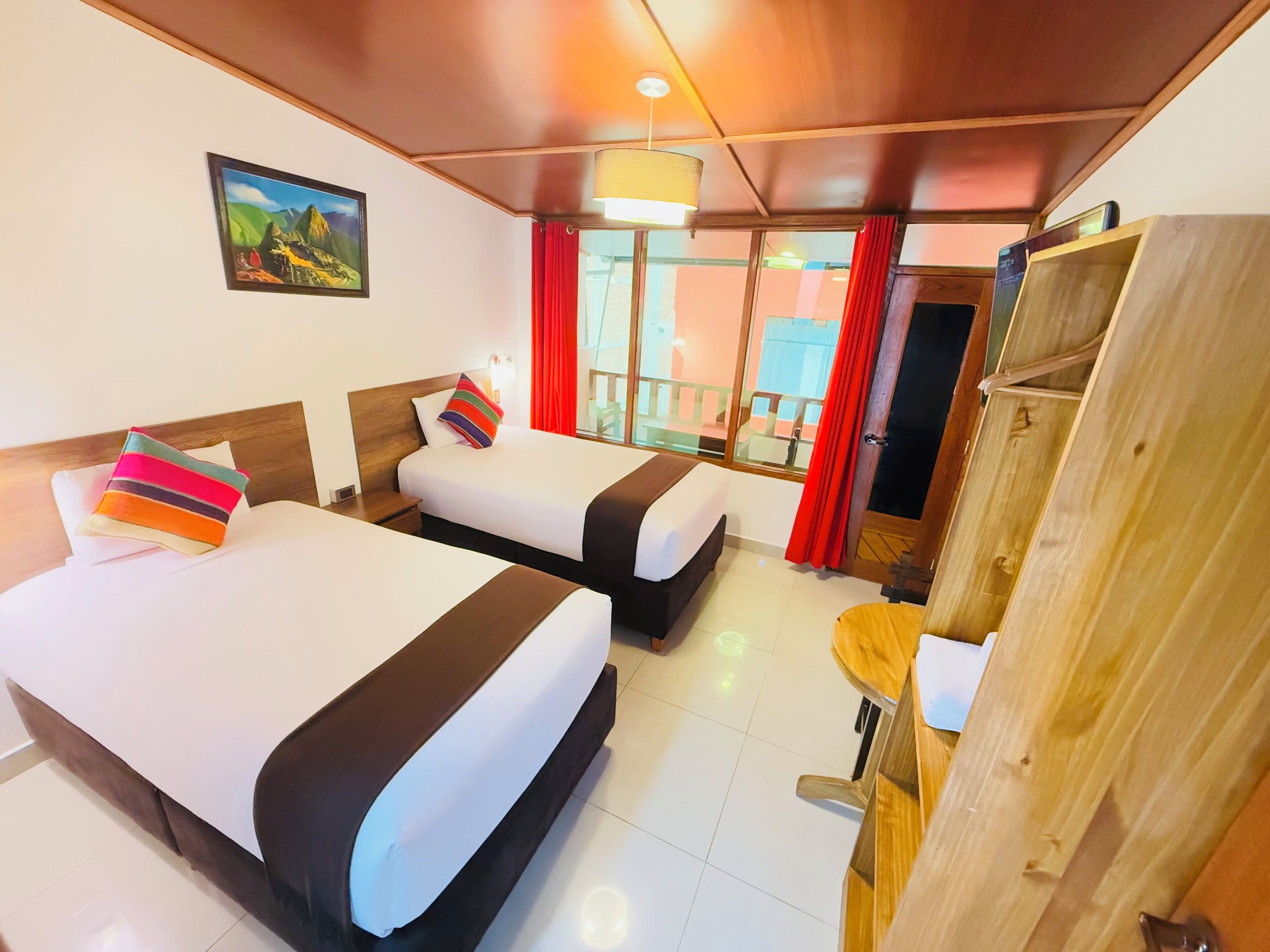 A hotel room with two beds, a wooden wardrobe, a small table, and a window with red curtains.