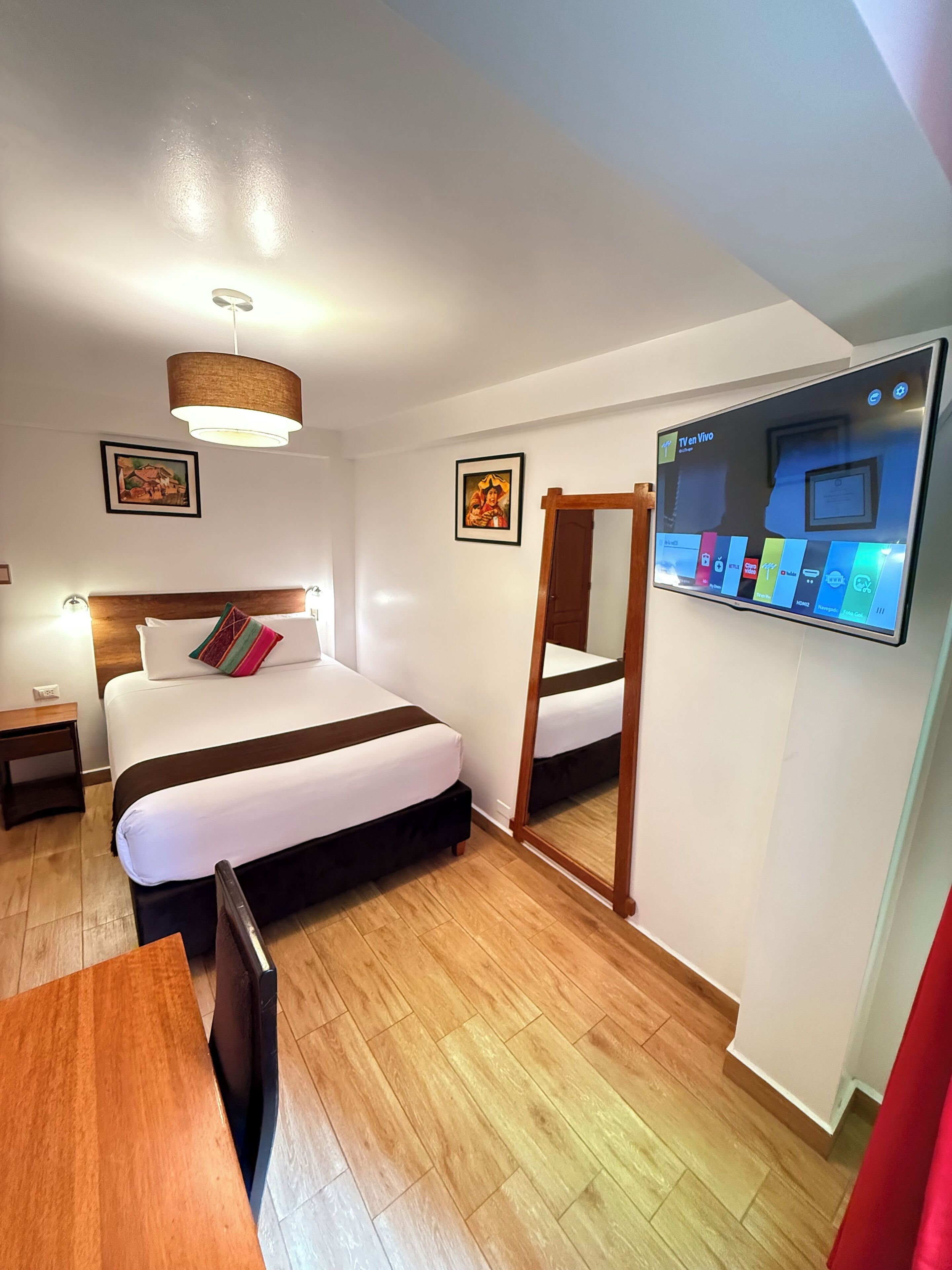 A hotel room with a bed, bedside table, lamp, TV, and wooden floor.