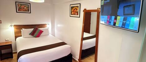 A hotel room with a bed, bedside table, lamp, TV, and wooden floor.
