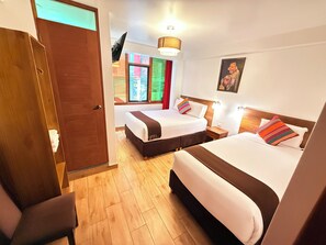A hotel room with two beds, wooden flooring, a painting on the wall, and a window with colorful curtains.