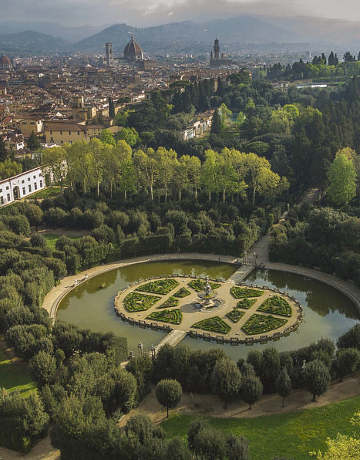 Vista aérea de un paisaje urbano con un parque circular destacado, con una fuente, rodeado de árboles y un camino.