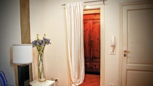 Um corredor com uma porta branca, uma porta de madeira, uma parede branca, uma cadeira azul, uma mesa branca com abajur e um vaso com flores.