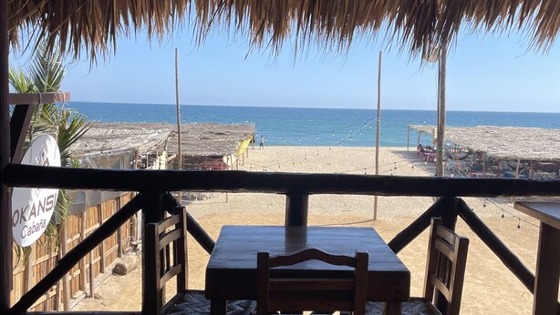 Een houten terras met rieten dak, uitkijkend op een strand.