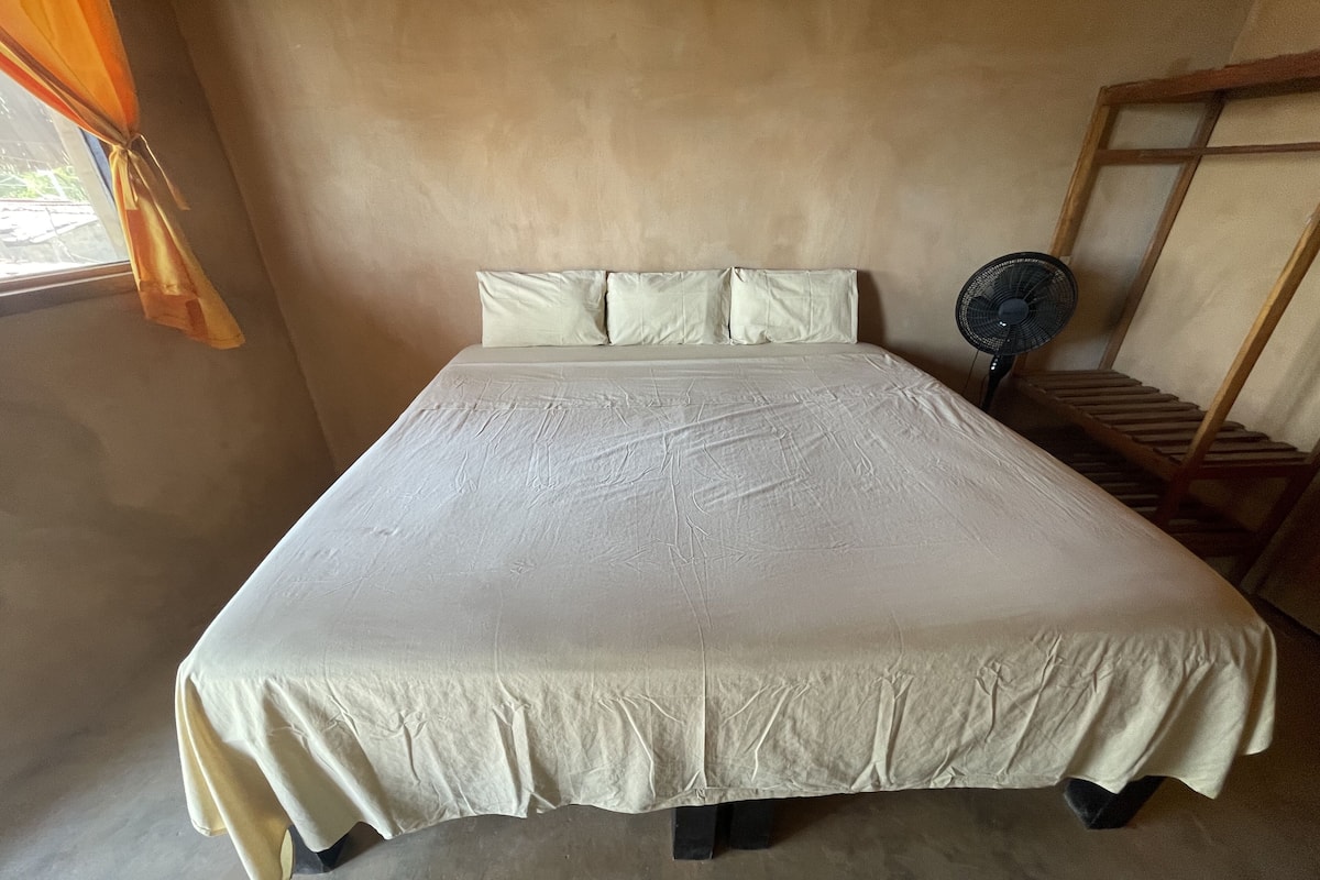 Un lit recouvert d’une courtepointe blanche et agrémenté d’oreillers, un ventilateur sur pied et une chaise en bois, le tout placé contre un mur de couleur beige.