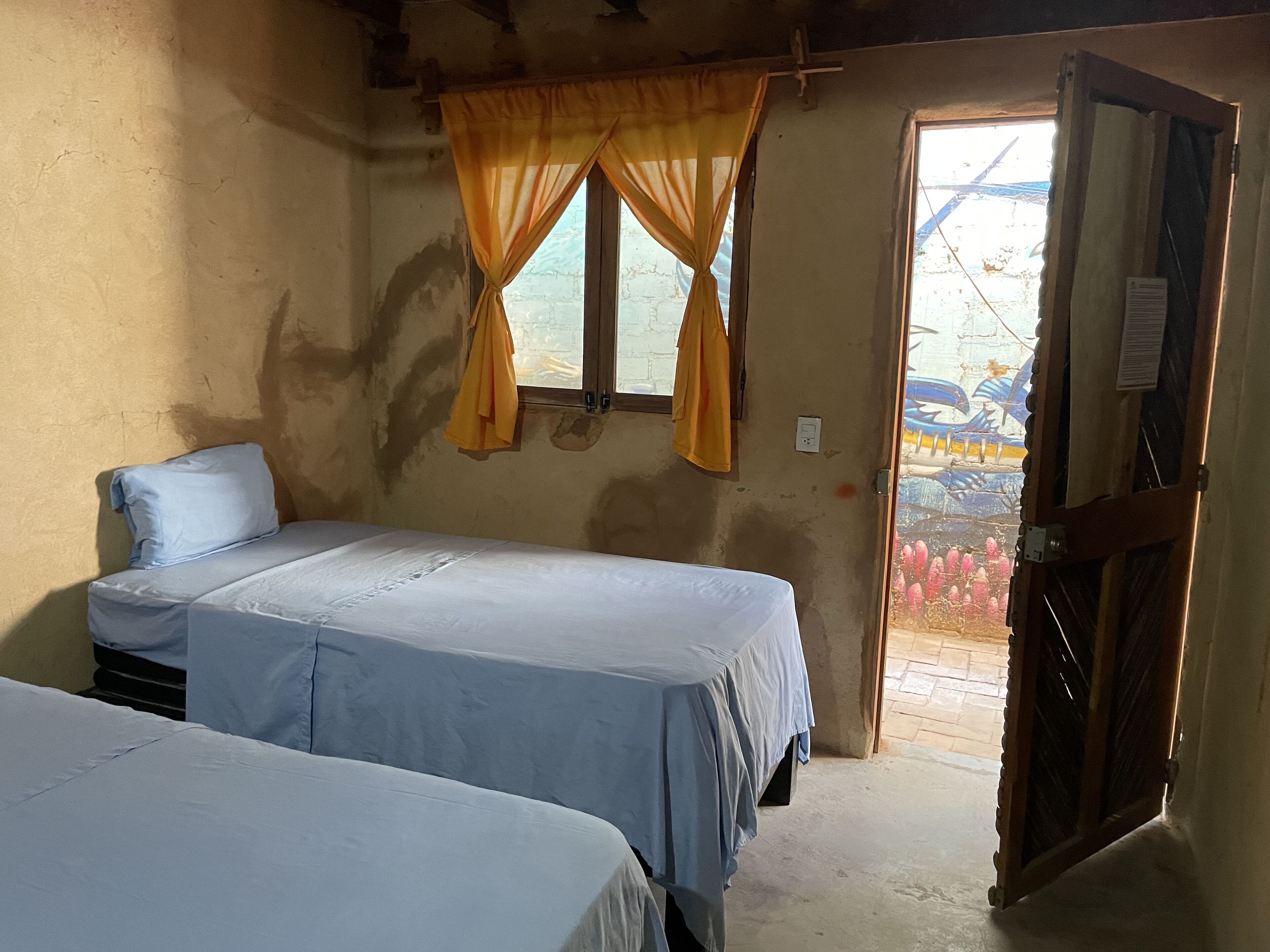 Una habitación con dos camas, una ventana con cortinas amarillas y una puerta que da al exterior.