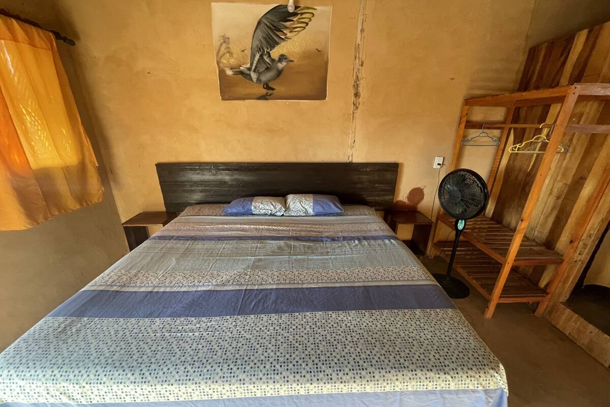 Une chambre à coucher avec un lit, une tête de lit en bois, un ventilateur fixé au mur et une étagère en bois.
