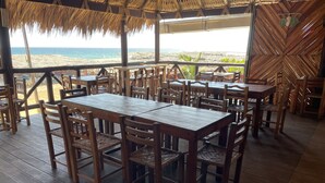 Een eethoek buiten met houten tafels en stoelen, met uitzicht op een strand.