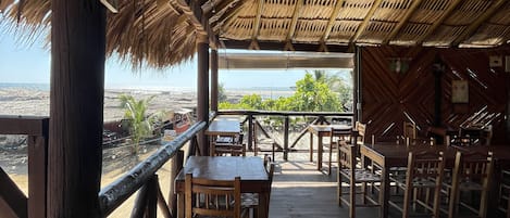 Un área de comedor al aire libre con mesas y sillas de madera, techo de paja y vista al océano.