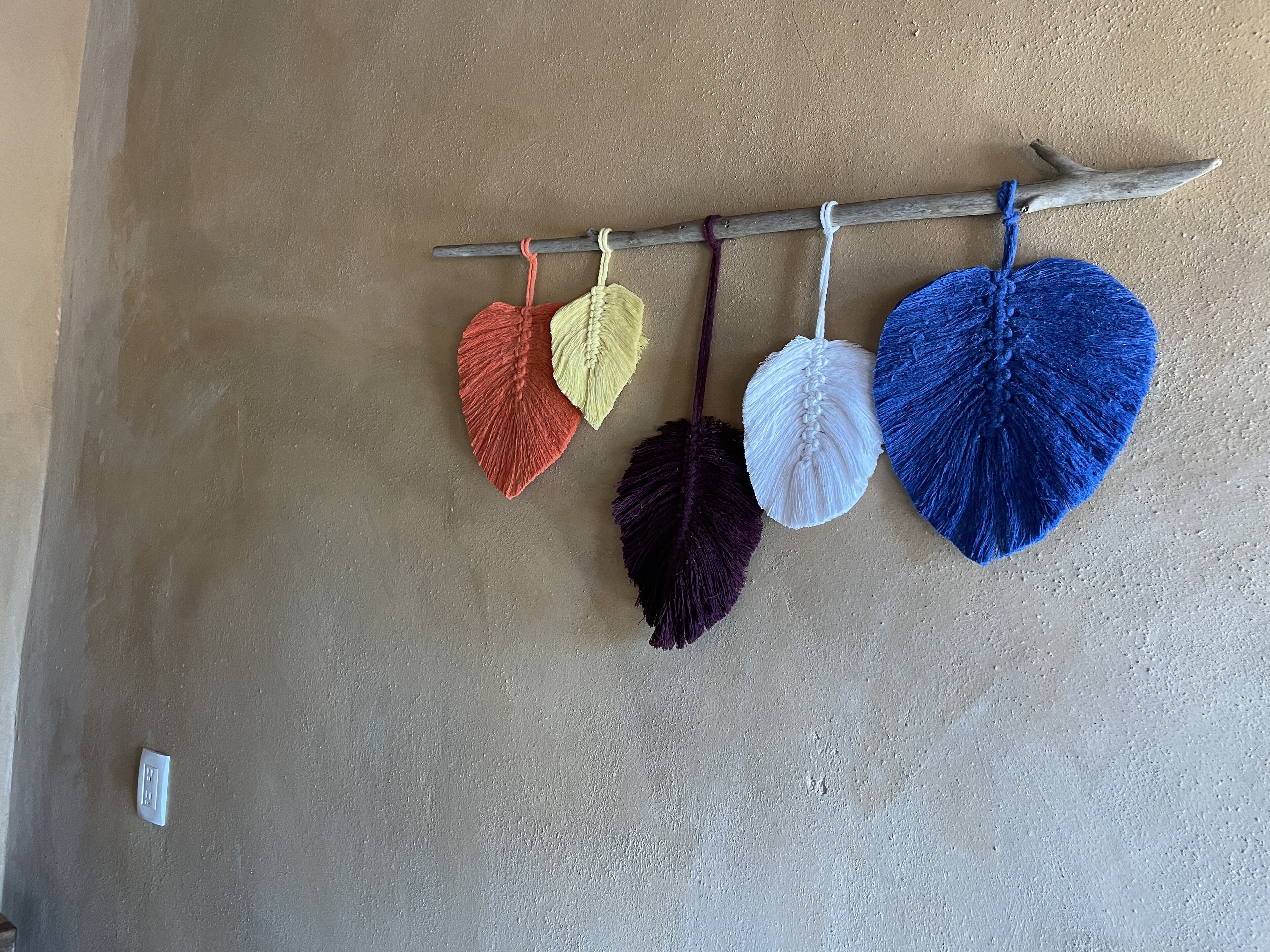 Un mur à la surface texturée et une branche en bois ornée de décorations en macramé aux couleurs vives.