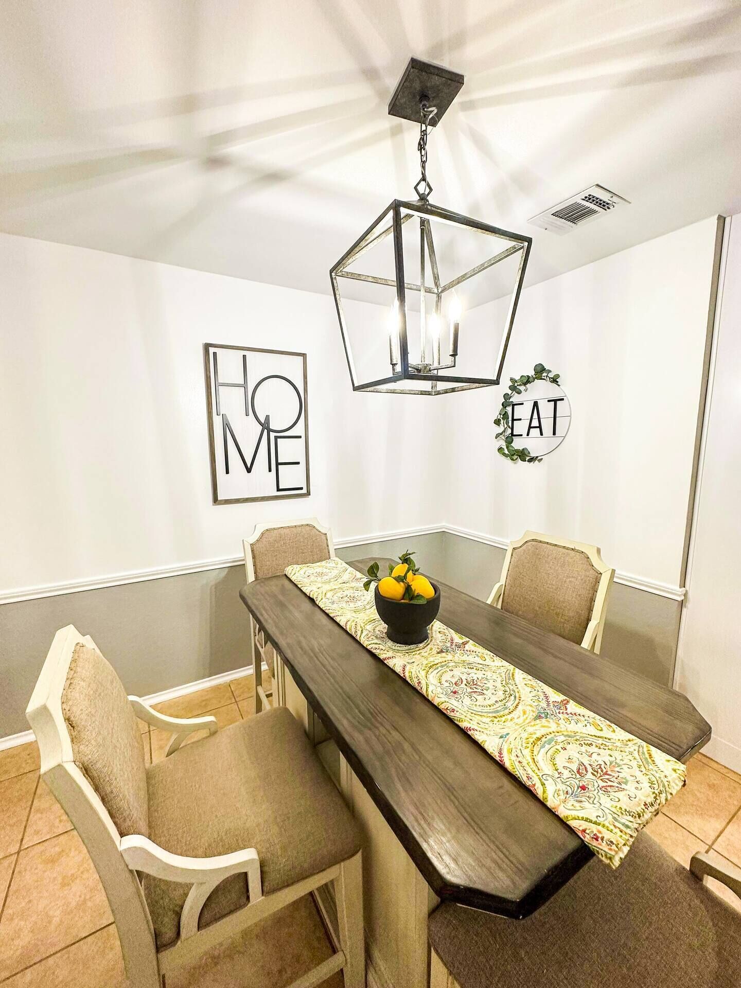 Una zona de menjador amb taula de fusta, cadires beix, corredor de taula estampat, lampadari modern i obra d'art mural amb la paraula “HOME” i “EAT”.