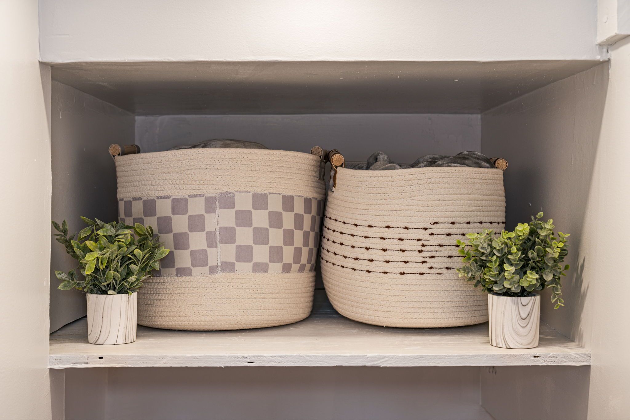 Two woven baskets on a shelf, each with a checkered pattern and a small potted plant beside them.