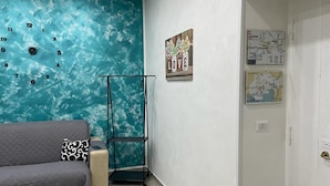 A room with a teal wall clock, a sofa, a metal stand, and two framed pictures. - Tutta nata storia (Naples)