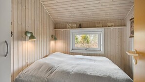 Makuuhuoneessa on sänky, kaksi seinään kiinnitettyä lamppua, ikkuna, josta on näkymä puita kohti, ja puupanelointi.