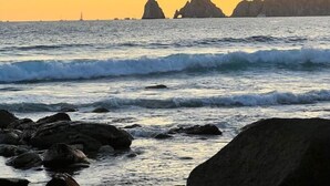 Paesaggio costiero con onde, rocce e montagne lontane sotto un cielo al tramonto.