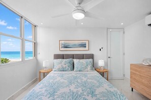 Een slaapkamer met een bed, twee nachtkastjes, een plafondventilator, een ingelijste foto en uitzicht op de oceaan.