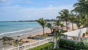 A beach view with a white fence, palm trees, and a small structure near the water.