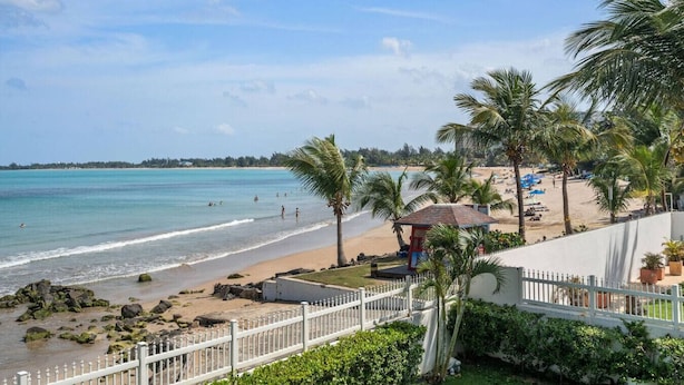 A beach view with a white fence, palm trees, and a small structure near the water. - Beachfront Sanctuary Newly Renovated to Perfection with Full Power Generator (Carolina)